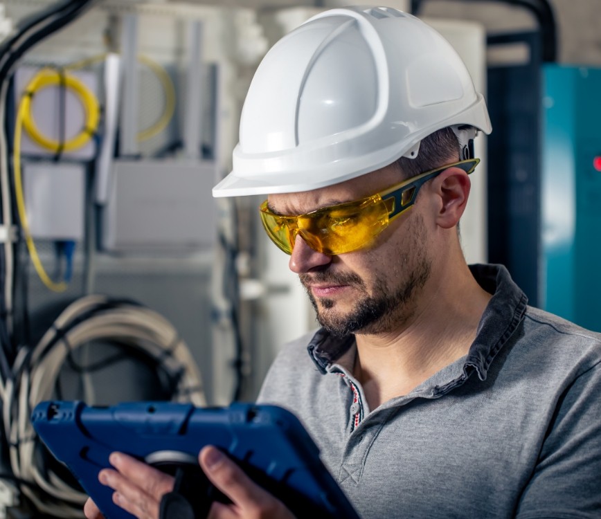 Técnico eléctrico con casco y gafas de seguridad inspeccionando datos en una tablet en una sala de control o instalación industrial