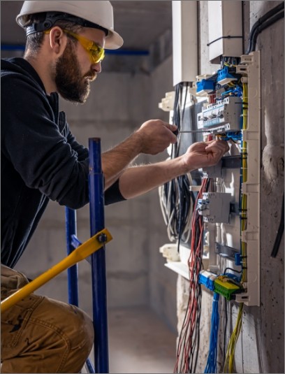 Técnico electricista con casco y gafas de seguridad realizando mantenimiento en un tablero eléctrico dentro de una construcción.